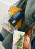 Woman holding two black net bags called Ida over her shoulder. Bags are full of newspapers, books, glasses & bread. 